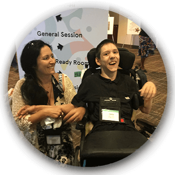 canchild-main A woman and a man in a wheelchair smile together at an indoor event; a sign behind them lists session locations.
