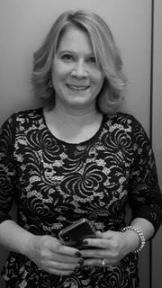Anne M A woman with shoulder-length hair smiles while holding a smartphone. She is wearing a patterned top and a bracelet. The image is in black and white.