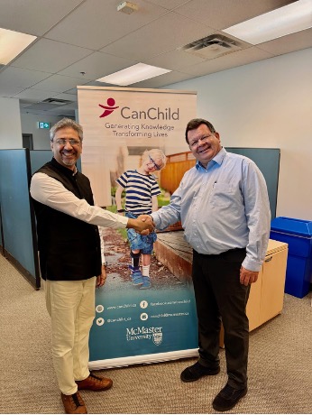 Two men shake hands in front of a CanChild banner at an office. The banner shows a child and reads "Generating Knowledge, Transforming Lives" with McMaster University branding.