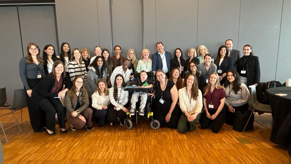 A large group of people poses together indoors, with several seated, some standing, and one person in a wheelchair at the center; professional attire, conference badges visible.