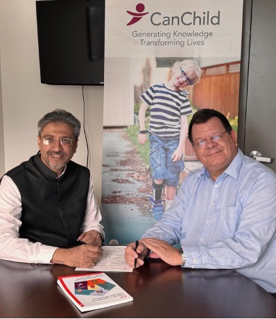 Two men sit at a table signing documents, with a CanChild banner featuring a child in the background and a book on the table.