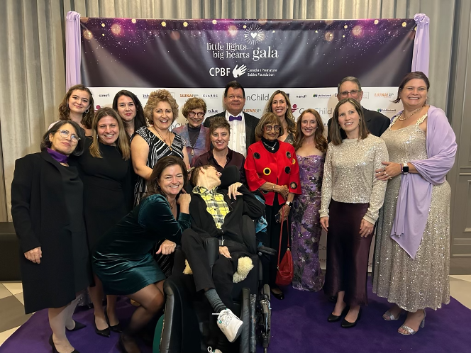 A group of people pose and smile together on the purple carpet at the Little Lights, Big Hearts Gala, with a branded backdrop behind them.