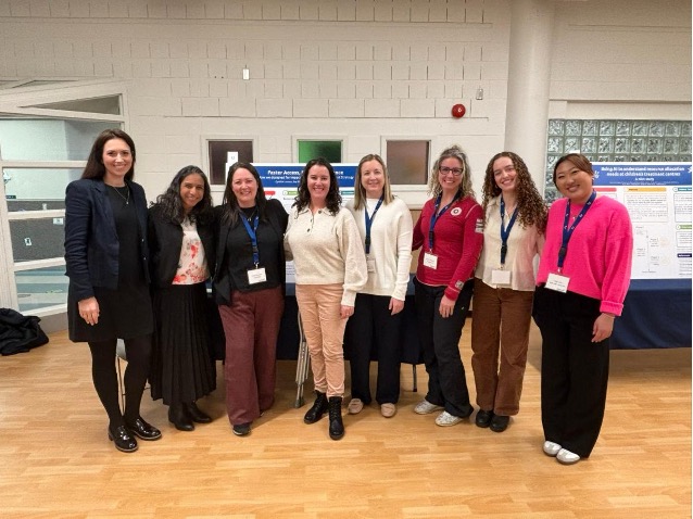 Eight women stand in a row indoors, all wearing conference badges, in front of research posters on display.