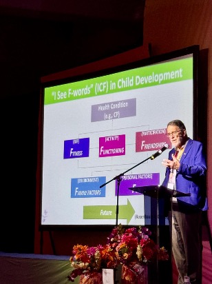 A speaker in a blue suit presents a slide titled "'I See F-words' (ICF) in Child Development" at a conference, with a podium and flower arrangement visible.