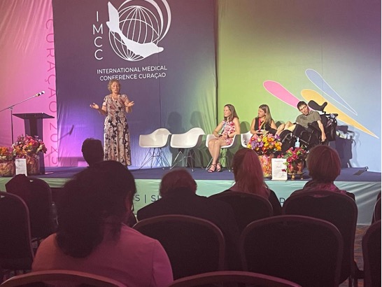 A woman speaks on stage at the International Medical Conference Curaçao, with three panelists seated beside her and an audience in the foreground.