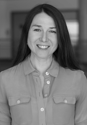 A woman with long dark hair, wearing a button-up shirt, smiles at the camera in a black and white photo.